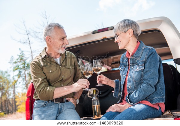 happy senior couple clinking wine glasses and looking\
at each other near car