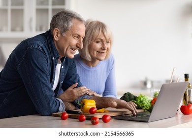 Happy Senior Couple Cooking Together At Home, Cheerful Elderly Man And Woman Reading Food Blog On Internet, Using Modern Laptop, Making Healthy Dinner, Kitchen Interior, Copy Space