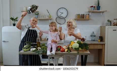 Happy Senior Couple Dancing With Granddaughter Child While Cooking In Kitchen At Home. Elderly Grandmother And Grandfather Making A Funny Dance With Strainers And Vegetables. Healthy Mature Family