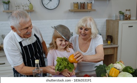 Happy Senior Couple Dancing With Granddaughter Child While Cooking In Kitchen At Home. Elderly Grandmother And Grandfather Making A Funny Dance With Strainers And Vegetables. Healthy Mature Family