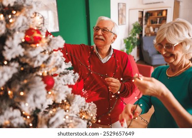 Happy senior couple decorating Christmas tree placing Christmas lights on it and having fun at home during winter holiday season - Powered by Shutterstock