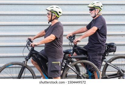 Happy Senior Couple Doing Together Outdoor Activity With Electric Bicycle. Healthy Lifestyle. Yellow Helmet, Black Clothes. Outdoor Against A Gray Metal Panel