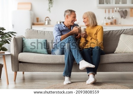 Happy Senior Couple Drinking Coffee Holding Mugs With Hot Drink And Smiling To Each Other Sitting On Couch At Home. On Weekend. Retirement Lifestyle And Happy Marriage Concept