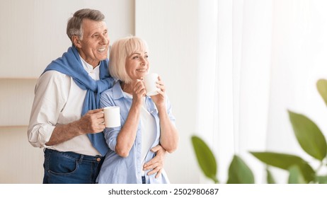 Happy senior couple drinking tea near window and embracing, free space - Powered by Shutterstock