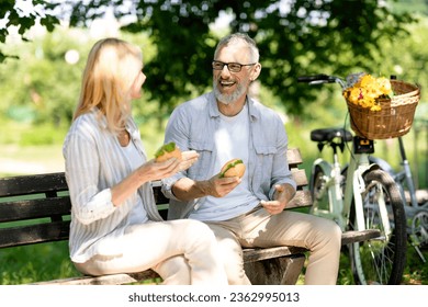 Happy Senior Couple Eating Sandwiches While Sitting On Bench In Park, Romantic Mature Spouses Having Lunch Outdoors And Laughing Together, Cheerful Older Man And Woman Enjoying Picnic Outside - Powered by Shutterstock