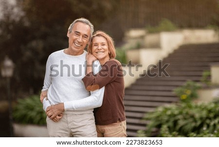 Happy Senior Couple Embracing Smiling To Camera Posing Standing In Green Park Outdoors, Wearing Casual Clothes. Love Relationship And Happiness In Mature Age. Retirement Lifestyle Concept