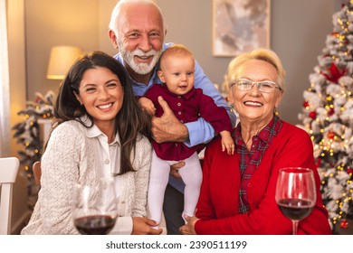 Happy senior couple enjoying spending Christmas with daughter and granddaughter, gathered around the table for Christmas dinner, grandfather holding baby girl - Powered by Shutterstock