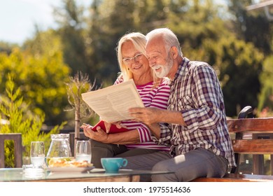 Happy senior couple enjoying their time together having an outdoor breakfast in the backyard of their home, man reading newspaper while woman is reading a book