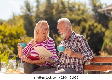 Happy senior couple enjoying their time together drinking coffee in the backyard of their home, man reading newspaper while woman is reading a book - Powered by Shutterstock