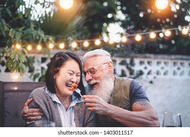 Happy senior couple having fun at dinner house party - Older people with different ethnicity doing a romantic date for celebrating anniversary - Elderly, food, drink and love concept