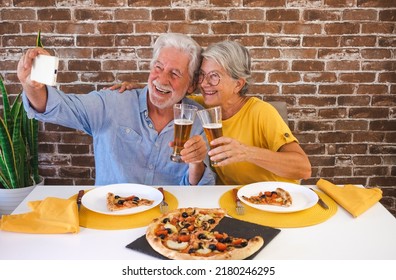 Happy Senior Couple Having Fun Together With Pizza And Beer. Elderly Woman And Man Laughing Sitting At Home Table Using Phone For A Selfie