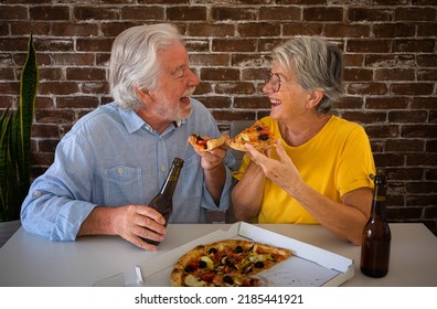 Happy Senior Couple Having Fun Together Eating A Pizza. Elderly Woman And Man Laughing Sitting At Home Table With Beer And Pizza