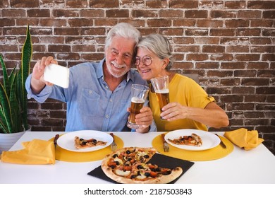 Happy Senior Couple Having Fun Together With Pizza And Beer. Elderly Woman And Man Laughing Sitting At Home Table Using Phone For A Selfie