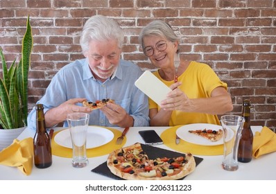 Happy Senior Couple Having Fun Together With Pizza And Beer. Elderly Woman And Man Laughing Sitting At Home Table Using Phone For A Selfie