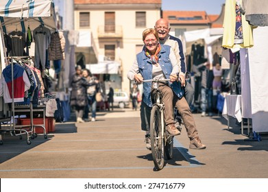 Happy Senior Couple Having Fun With Bicycle At Flea Market - Concept Of Active Playful Elderly With Bike During Retirement - Everyday Joy Lifestyle Without Age Limitation In A Spring Sunny Afternoon