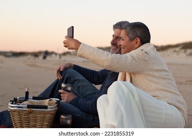 Happy senior couple having picnic at seashore in evening, drinking wine and taking selfie on mobile phone. Grey-haired man smiling while lady holding smartphone. Modern technology, romance concept - Powered by Shutterstock