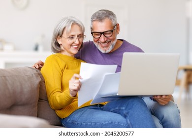 Happy Senior Couple Holding Financial Documents And Reading Good News In Letter, Smiling Mature Family Paying Bills Online On Laptop While Sitting On Sofa At Home, Discussing Money Income