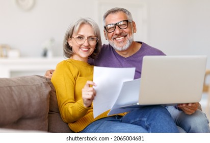 Happy Senior Couple Holding Financial Documents And Reading Good News In Letter, Smiling Mature Family Paying Bills Online On Laptop While Sitting On Sofa At Home, Discussing Money Income