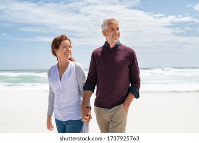 Happy Senior Couple Holding Hands At Beach In A Sunny Autumn Day. Loving Mature Couple Enjoying Their Vacation At The Sea Shore With Copy Space. Smiling Wife And Happy Old Husband Walking Barefoot.