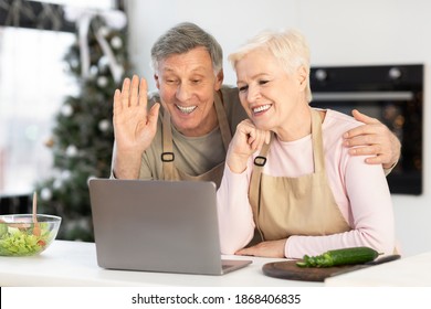 Happy Senior Couple At Laptop Making Video Call In Kitchen Celebrating Christmas Holiday Remotely. Mature Spouses Cooking Preparing Xmas Dinner And Congratulating Distant Family On New Year Online