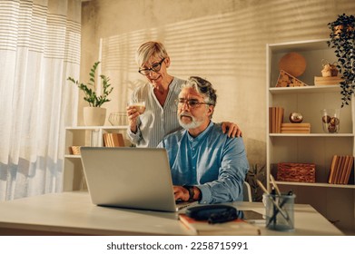 Happy senior couple laughing and bonding while using laptop at home. Smiling elderly husband and wife having fun while browsing internet or reading a good news. Copy space. Drinking coffee. - Powered by Shutterstock