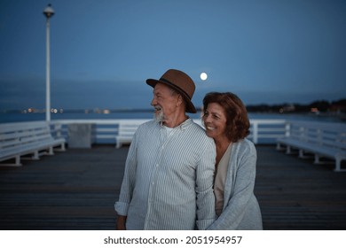 Happy Senior Couple In Love On Walk Holding Hands Outdoors On Pier By Sea At Moonlight, Looking At View.