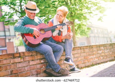 Happy Senior Couple Playing A Guitar While Sitting Outside On A Wall On A Sunny Day - Concept Of Active Elderly Having Fun With Guitar - Enjoying Lifestyle During Retirement