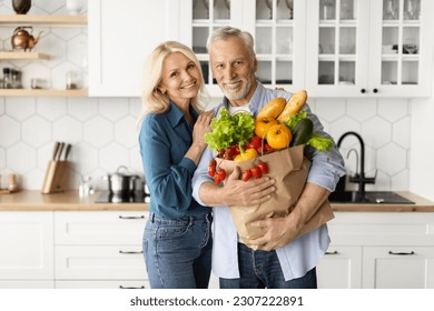 Happy senior couple posing with grocery bag in kitchen interior, elderly retired spouses holding pack with fresh vegetables at home, looking and smiling at camera, enjoying life on retirement - Powered by Shutterstock