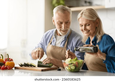 Happy Senior Couple Preparing Tasty Lunch Together In Kitchen, Smiling Mature Man And Woman Cooking Healthy Vegetarian Meal At Home, Woman Adding Oil To Fresh Vegetable Salad, Free Space - Powered by Shutterstock