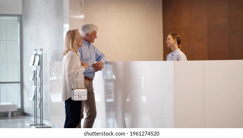 Happy Senior Couple Receiving Tourist Information At Hotel Reception. Mature Elegant Man And Woman Talking To Concierge In Apartment House Lobby