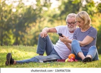 Happy Senior Couple Relaxing In Park Using Smartphone Together . Old People Sitting On Grass In The Summer Park Looking Mobile Phone . Elderly Resting .mature Relationships