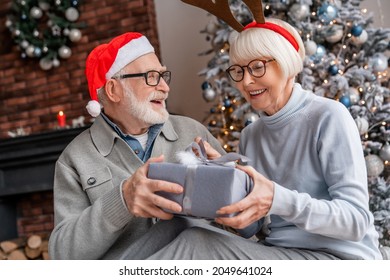 Happy senior couple in Santa hats exchanging Christmas gifts at home. Grandparents celebrating New Year together near tree fir decorated with ornaments. - Powered by Shutterstock