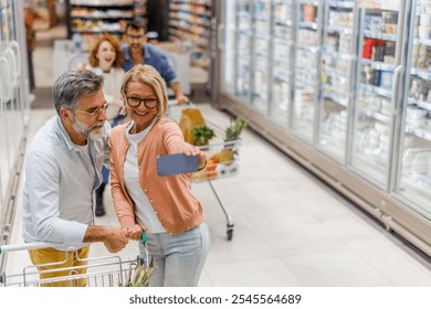 Happy senior couple shopping at the supermarket, taking a selfie while browsing groceries. Smiling and pushing a cart filled with produce, embracing a healthy lifestyle with modern technology - Powered by Shutterstock