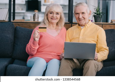 Happy Senior Couple Sitting On Couch With Laptop And Credit Card, Looking At Camera And Doing Online Shopping At Home