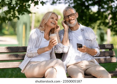 Happy senior couple sitting on park benchand listening music in earphones, cheerful mature spouses using smartphone and drinking takeaway coffee, romantic older man and woman sharing headphones - Powered by Shutterstock