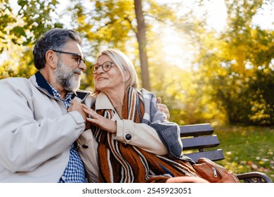 Happy senior couple sitting on a park bench in autumn, smiling and enjoying a sunny day outdoors with colorful leaves in the background. - Powered by Shutterstock