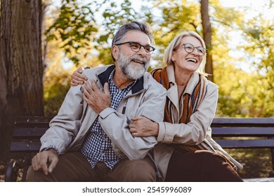 Happy senior couple sitting on a park bench in autumn, smiling and enjoying a sunny day outdoors with colorful leaves in the background. - Powered by Shutterstock