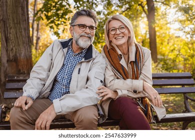 Happy senior couple sitting on a park bench in autumn, smiling and enjoying a sunny day outdoors with colorful leaves in the background. - Powered by Shutterstock