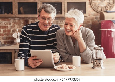 A happy senior couple are sitting in their kitchen, looking at a tablet computer. The woman has a hand on her chin and is laughing, while the man is holding the tablet and smiling - Powered by Shutterstock