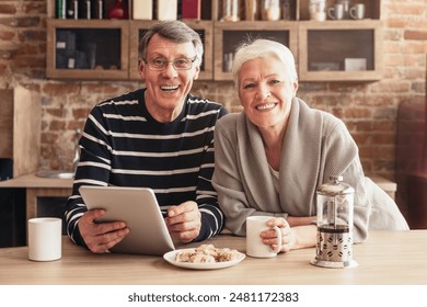 A happy senior couple smiles for the camera while sitting at a kitchen table, enjoying coffee and using a tablet. The man is wearing a striped shirt, and the woman is wearing a grey sweater. - Powered by Shutterstock