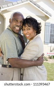 Happy Senior Couple Standing In Front Of House For Sale