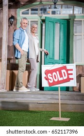 Happy Senior Couple Standing On Porch Of New House And Sold Sign On Green Grass