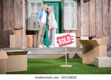 Happy Senior Couple Standing On Porch Of New House And Sold Sign On Green Grass