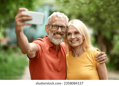 Happy Senior Couple Taking Selfie On Smartphone Outdoors, Cheerful Mature Spouses Capturing Photos While Standing In Garden, Embracing And Smiling At Mobile Phone Camera, Enjoying Time Together - Powered by Shutterstock