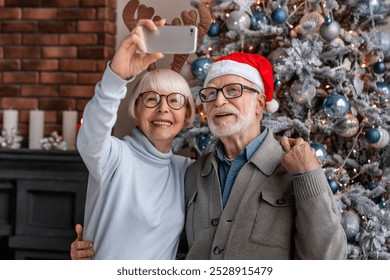 Happy senior couple taking selfie near decorated Christmas tree at living room. Caucasian old elderly couple spouses grandparents vlogging blogging having videocall on New Year Eve - Powered by Shutterstock