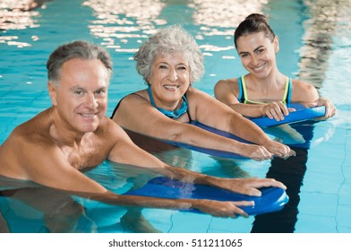Happy senior couple taking swimming lessons from young trainer. Smiling old woman and mature man doing aqua aerobics exercise in swimming pool. Retired people in swimming pool looking at camera.