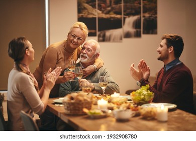 Happy senior couple toasting with wine while their adult children are applauding for their marriage anniversary at dining table.