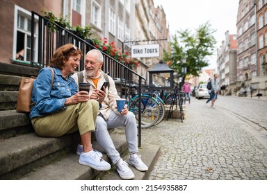 Happy senior couple tourists sitting on stairs and having take away coffee outdoors in town