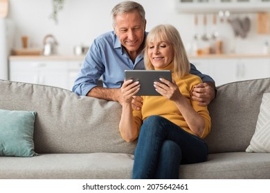 Happy Senior Couple Using Digital Tablet Together Browsing Internet Sitting On Sofa At Home. Retired Husband And Wife Reading Online News On Computer On Weekend. Mature People And Gadgets