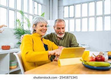 Happy senior couple using digital tablet at home pointing at the screen and smiling while eating breakfast together - Powered by Shutterstock